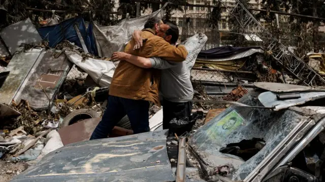 Two men hug in front of a destroyed car. Rubble from damaged buildings can be seen behind the pair