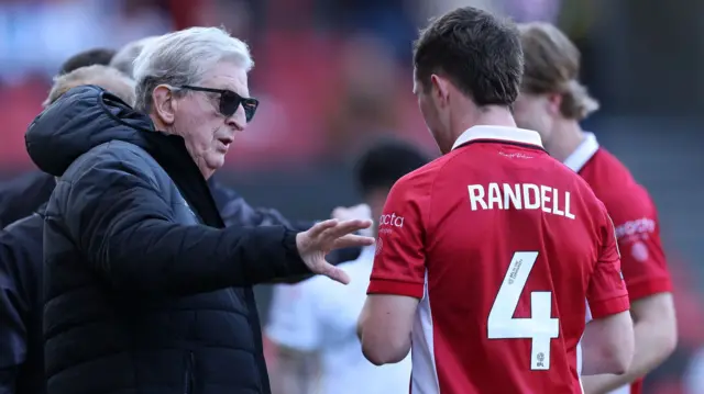 A photo of Roy Hodgson, wearing a black coat and sunglasses, speaking to Adam Randell