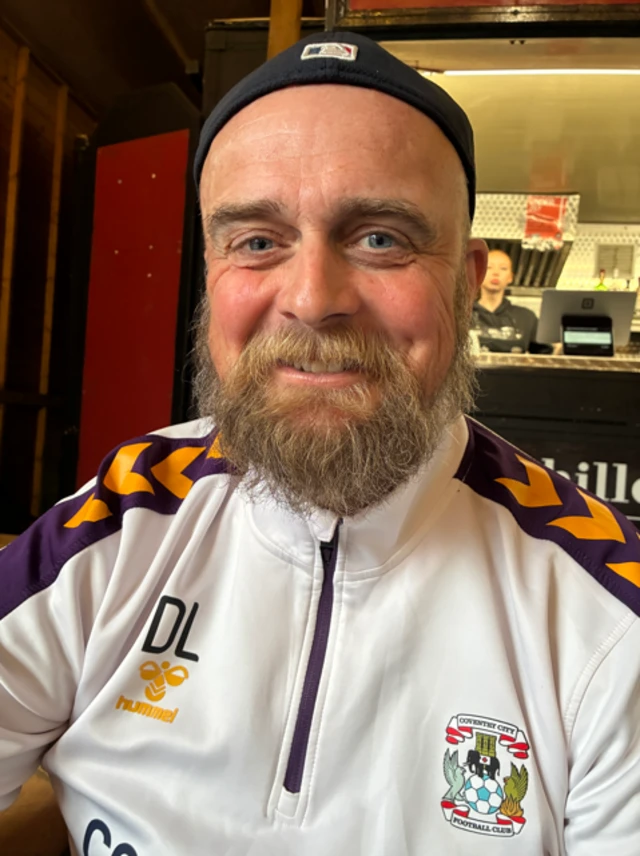 A man wearing a white Coventry City zip up top smiles at the camera - he is wearing a black beanie hat, has a moustache and beard and is smiling directly at the camera