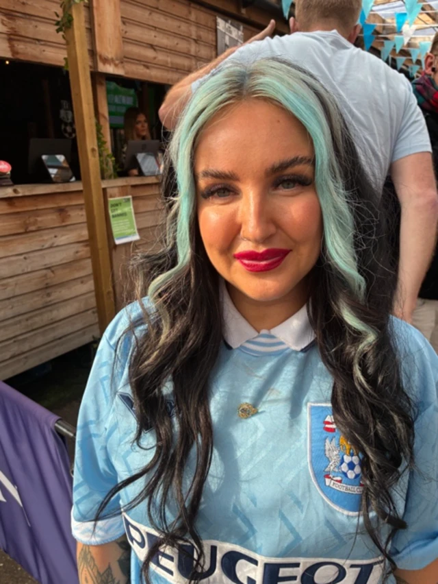 A woman with long dark curly hair with two light blue streaks at the side - she is wearing a Coventry City shirt