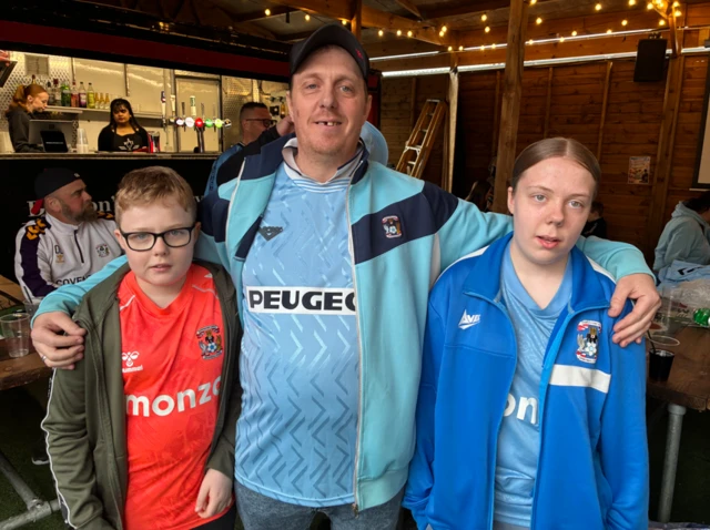 A man in a Coventry City shirt and jacket hugs his son and daughter who are both dressed in Coventry gear