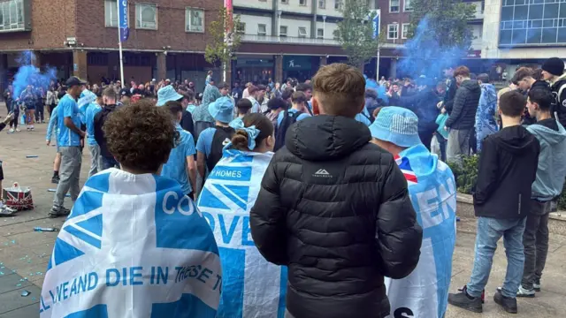 Young fans with flags around their shoulders and some blue flares can be seen.