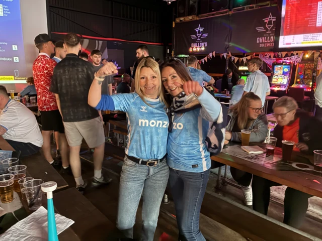 Two women in Coventry City shirts and jeans hug each other and give a thumbs up to the camera. They are in a pub and there is a crowd of people behind them
