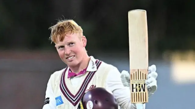 James Rew holding his bat up with his left arm and his helmet under his shoulder after celebrating a half century.