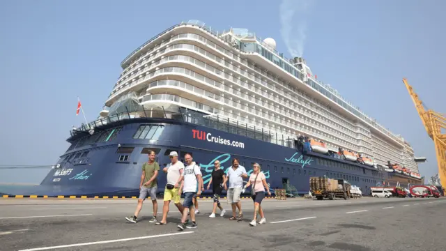 Tourists walk in front of huge cruise ship which is docked at port