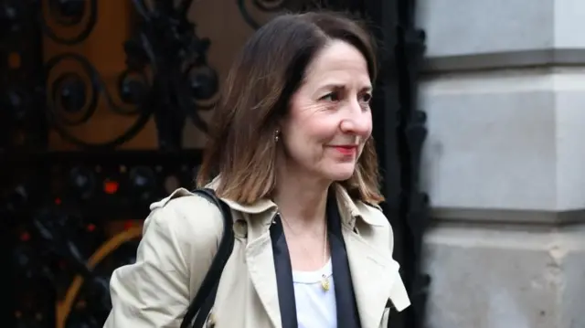 Liz Kendall, a woman with shoulder-length brown hair, wears a beige overcoat in front of a black gate