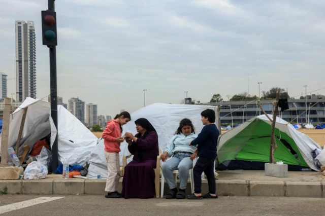 A woman with a black head covering veil and clothing sits on a plastic chair next to her eldest daughter as she looks at something in her hands with her youngest daughter. A boy stands next to the chair talking, behind the group are three tents pitched on a street