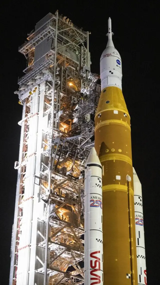 The Orion spacecraft, with orange and white colours, shown upright against a dark sky.