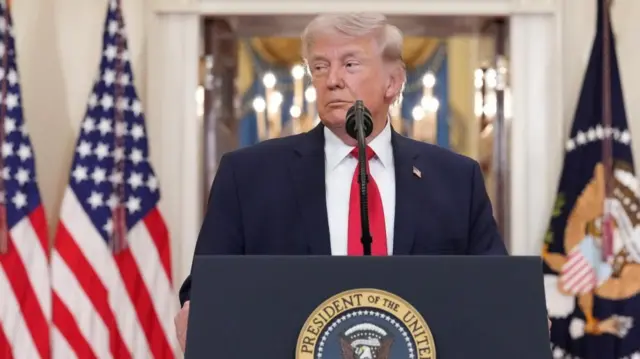 Donald Trump standing behind podium with US flags waving behind him