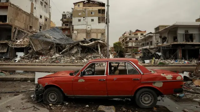 red car, slightly damaged, in front of collapsed and destroyed buildings