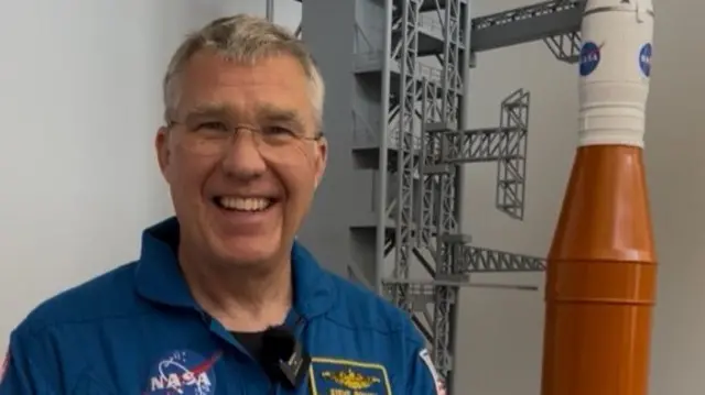 Stephen smiling in a blue jumpsuit standing next to a model rocket