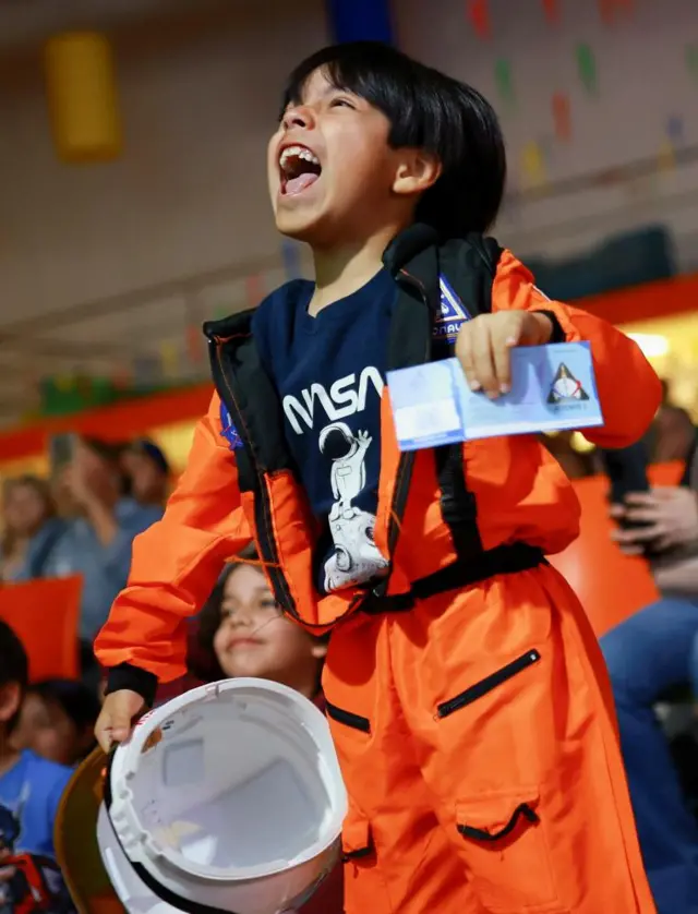 A child dressed in an orange space jumpsuit. he holds a white helmet under his arm and is smiling, cheering. he has medium length black hair.