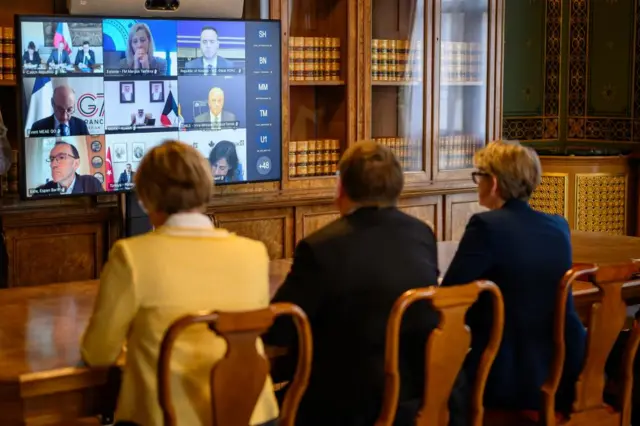 Three people sit facing a large TV screen displaying other members of a virtual meeting in an ornate panelled room