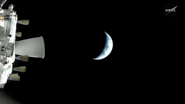 A partial sliver of the Earth, known as crescent Earth, is seen from the Orion spacecraft. Black surrounds the earth and the edge of the spacecraft is visible in the left of the frame.