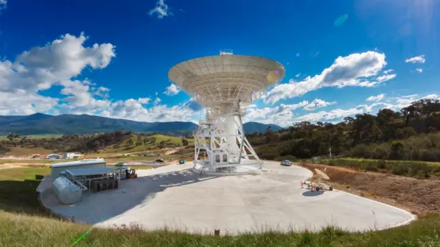 A giant satellite sits on concrete pad, surrounding is some brown grass and rolling hills in the background. The sky is blue with some clouds in the distance