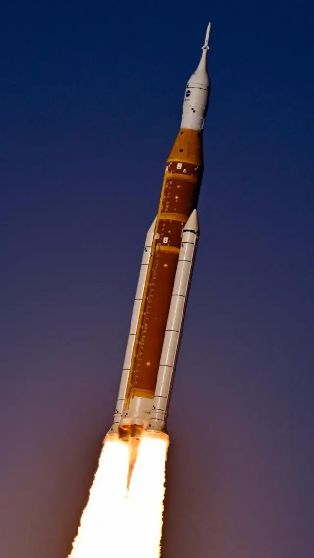 Artemis II is seen shortly after lift off from Cape Canaveral. The orange rocket body is flanked by two white propulsion rockets, with bright fire coming from beneath them. a dark blue sky is seen behind.
