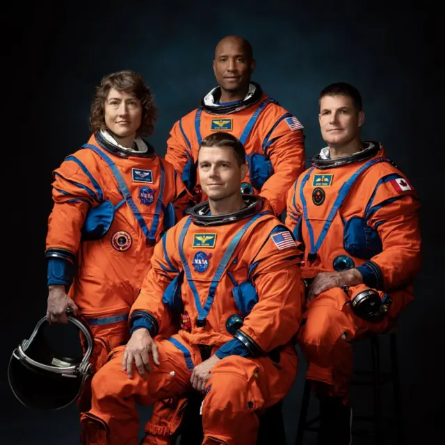 Four astronauts are seen in their orange spacesuits arranged for a professional picture. The suits have blue detailing and they are positioned in front of a dark blue background.