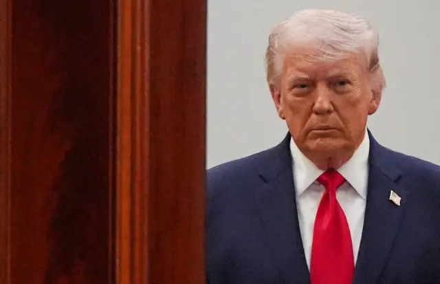 Donald Trump in dark blue suit, white shirt and red tie, with a US flag pin at the top of his left lapel, waits outside a wooden door