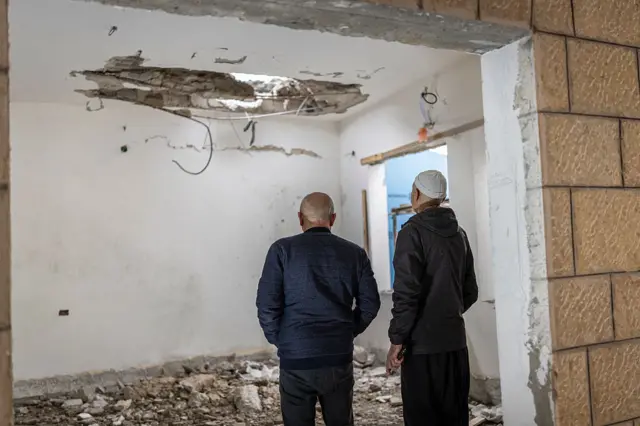 Two men in dark clothing stand inside a room of a residential building heavily damaged. A large hole in the ceiling, debris scattered across the floor