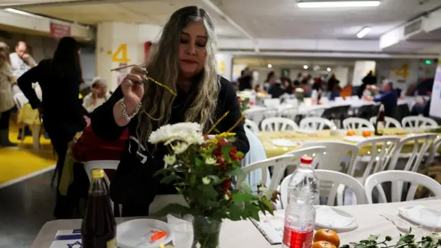 A woman arranging a flower bouquet in an underground parking garage. Behind her are lots of tables with plates set out to have a meal. There are lots of people gathered in the background.
