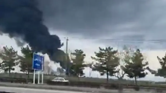 A screengrab from a video being filmed as a car drives by a large plume of black smoke rising into the air