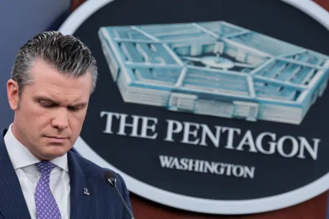 Pete Hegseth stands in front of a large seal of the Pentagon and looks down