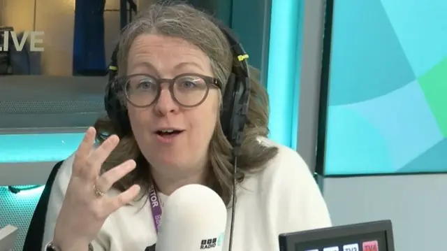Libby Jackson pictured at the 5Live studio in front of a microphone. She is wearing a cream-coloured top and wearing a purple 'visitor' lanyard.