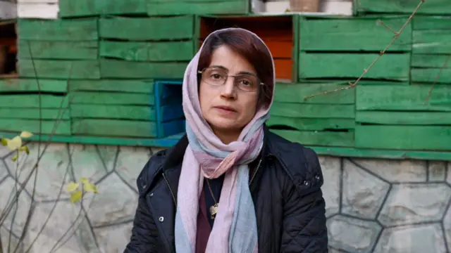 A woman wearing a headscarf stands in her garden, looking directly at the camera
