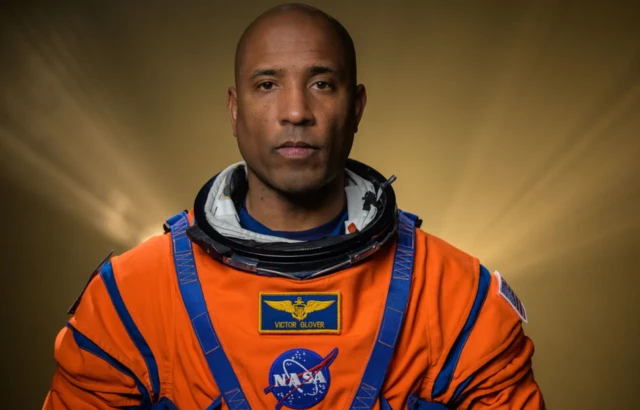 Victor Glover stands facing the camera in his orange space suit, there is a yellow background and rays of light blocked by his body