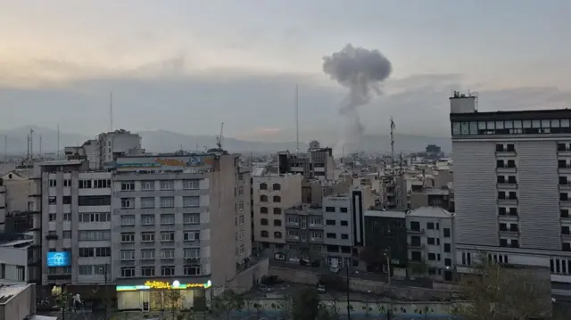 Wide shot of a residential area of Tehran, a grey cloud of smoke billowing into the air in the far background following an explosion
