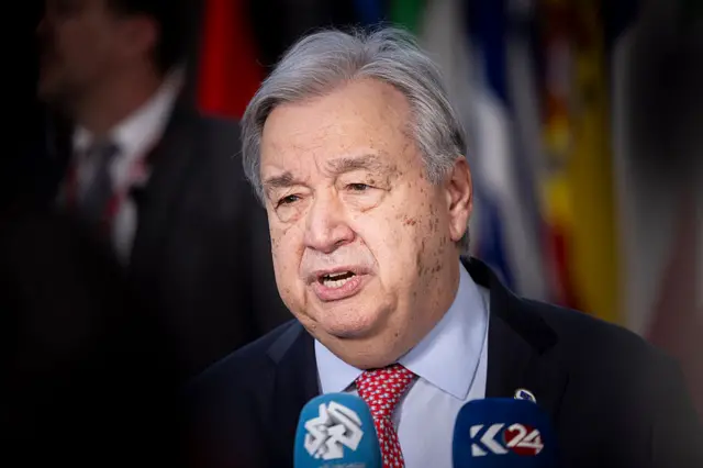 UN Secretary General António Guterres speaking with two microphones in front of him. He wears a black jacket and red patterned tie