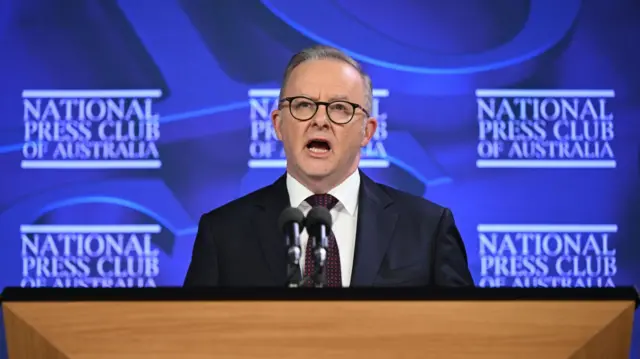 Anthony Albanese stands at a podium before a blue backdrop.