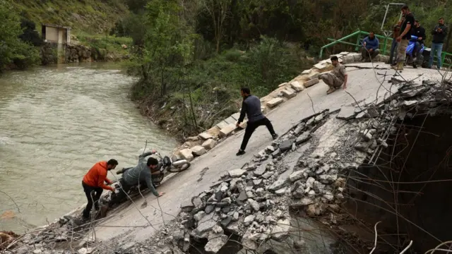 Algumas pessoas empurram uma motocicleta para cima de uma ponte danificada, com vários homens em pé no topo e quatro pessoas posicionadas na parte inclinada e danificada.