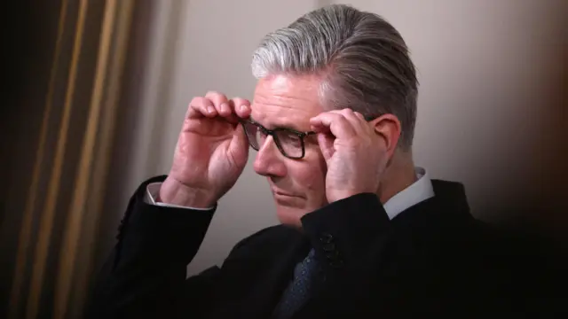 Sir Keir Starmer has short grey hair neatly combed and is wearing a black coat with dark blue tie and white shirt. His hands are adjusting his glasses and he looks serious