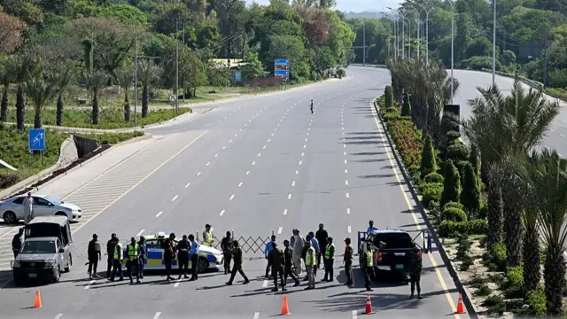 Security personnel are seen standing beside vehicles on an empty road. Trees line the pavement on either side of the road and orange traffic cones can be seen.