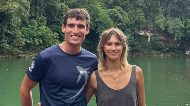 Sophie and Danny smiling with their arms around each other facing the camera. They stand against a black railing in front of green water, which is surrounded by green vegetation
