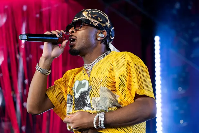A man wearing a yellow mesh top, sunglasses and a bandana on his head sings into a microphone