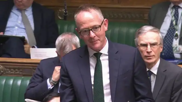 John Lamont speaks in Parliament, wearing a blue suit and green tie.