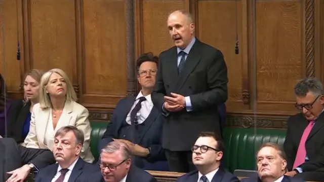 Simon Hoare speaks in Parliament, wearing a suit and tie.