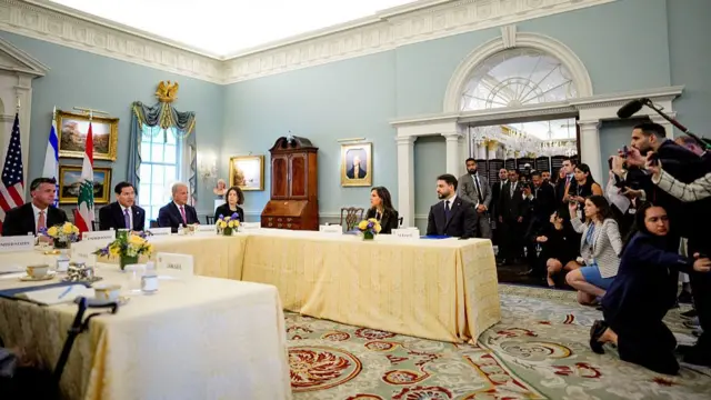 Several people sit around a U shaped table as several media representatives look on.