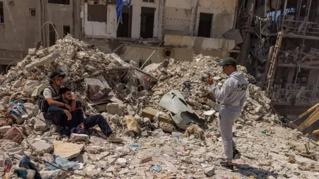 A man in a tracksuit and a cap takes a photograph on a phone of two men sitting in a pile of rubble among buildings attacked by strikes