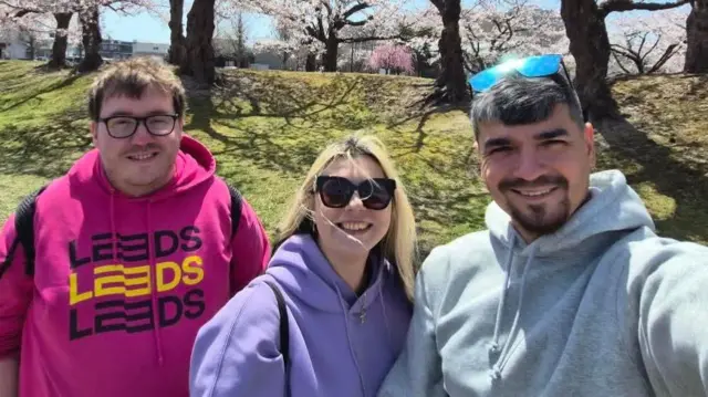 David stood smiling with a man and a woman in front of a line of blossom trees