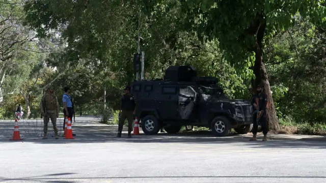 An armed black car parked on a road which is blocked by traffic cones and barbed wire. On the road stand army officers and police holding large guns
