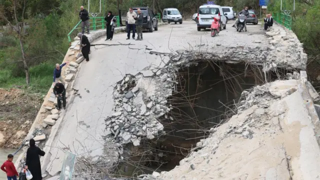 Uma grande abertura é visível em uma ponte danificada, com carros estacionados em cima dela. Várias pessoas estão posicionadas ao longo da estrutura.