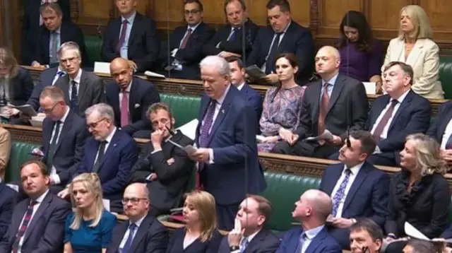 A group of MPs sitting on green sofas while Lewis stand up and speaks, reading off paper