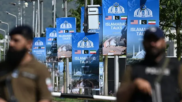 Blurred out security personnel are pictured in front of posters advertising talks between the US and Iran in Islamabad, with the posters being in focus.