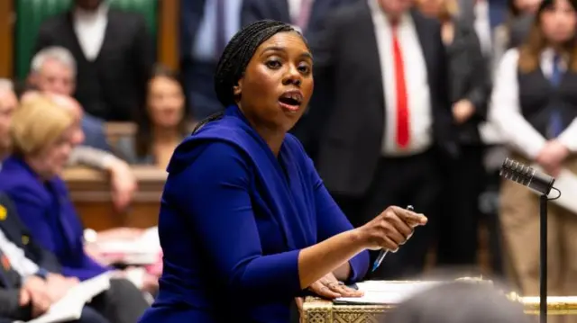 Kemi Badenoch speaks in Parliament. She is wearing a bright blue outfit and gesturing with her right hand.