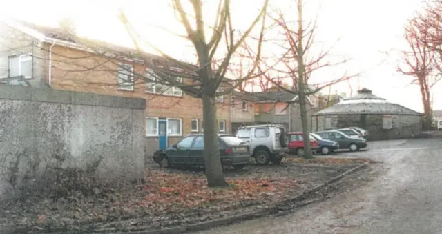 Exterior shot of a care home. Cars are parked in a row outside. Trees in the shot have dropped their leaves on the ground.