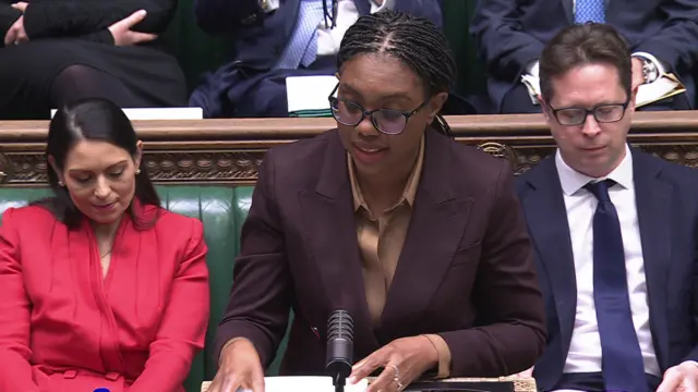 Kemi Badenoch standing in the House of Commons, wearing a dark brown blazer and light brown blouse