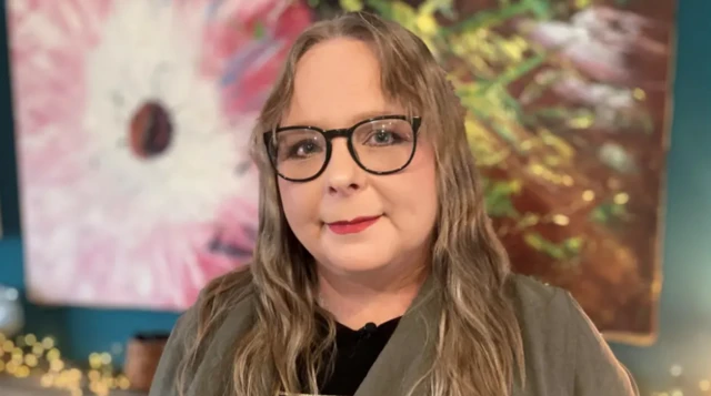 A woman with glasses and long, fair hair looks at the camera.